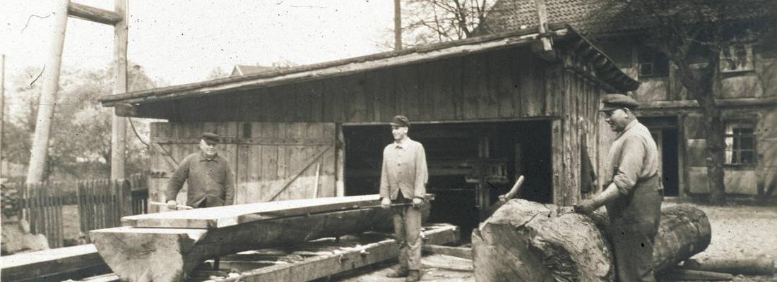 Drei Männer arbeiten mit Holz. Vor einem der Männer liegt ein großer Baumstamm, zwei anderer stehen neben einem gesägten Brett.
