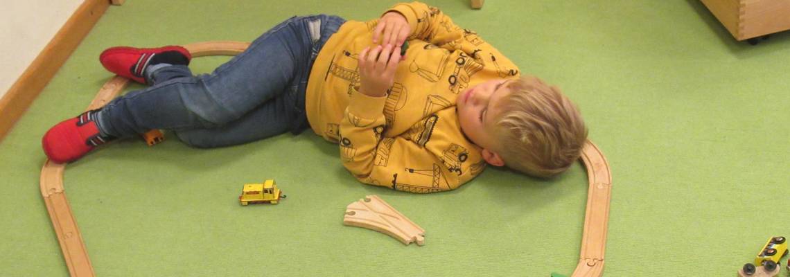 Ein Kind liegt auf dem Boden. Um es herum ist eine Holzeisenbahn aufgebaut.