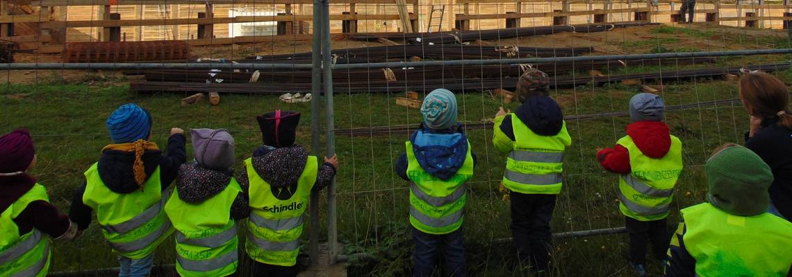 Eine Gruppe von Kindern steht an einem Bauzaun. Hinter dem Zaun sind Baufahrzeuge, alle Kinder tragen Warnwesten.