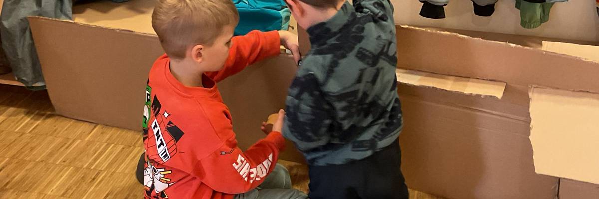 Zwei Jungs bauen in der Garderobe mit großen Pappkartons und Werkzeug. Sie knien gerade vor einem großen, länglichen Karton.