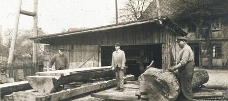 Drei Männer arbeiten mit Holz. Vor einem der Männer liegt ein großer Baumstamm, zwei anderer stehen neben einem gesägten Brett.