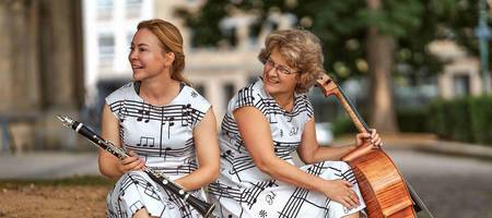 Zwei Frauen mit identischen Kleidern mit Musiknoten sitzen auf einem urbanen Pflanzbeet aus Beton. Die eine hält ihre Klarinette in der Hand, die andere ihr Cello.